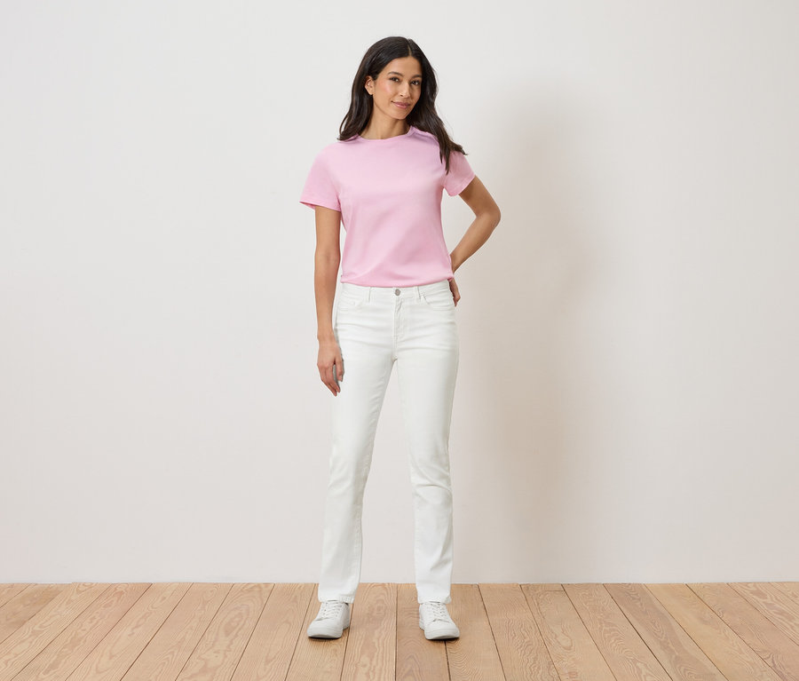 Žena stojí před bílou zdí a má na sobě růžové tričko, bílé džíny a bílé tenisky.