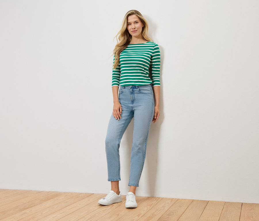 Blondýna stojí před zdí a má na sobě zeleno-bílé pruhované tričko s dlouhým rukávem, světle modré džíny a bílé tenisky.