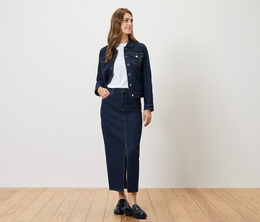 Žena stojí a má na sobě džínovou bundu a džínovou sukni s rozparkem.