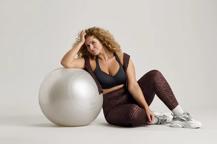 Žena s kudrnatými vlasy sedí vedle stříbrného gymnastického míče, má na sobě černou sportovní podprsenku, legíny se vzorem zebry a tenisky.