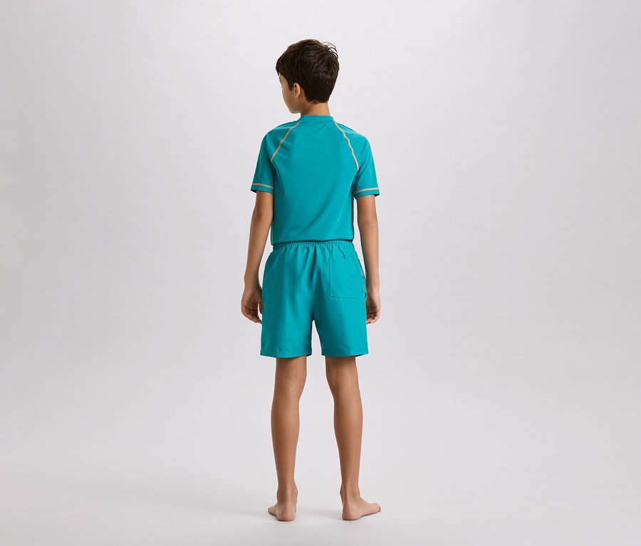 Chlapec zezadu, má na sobě tyrkysový set skládající se z topu a šortek se zlatými akcenty, na neutrálním pozadí.