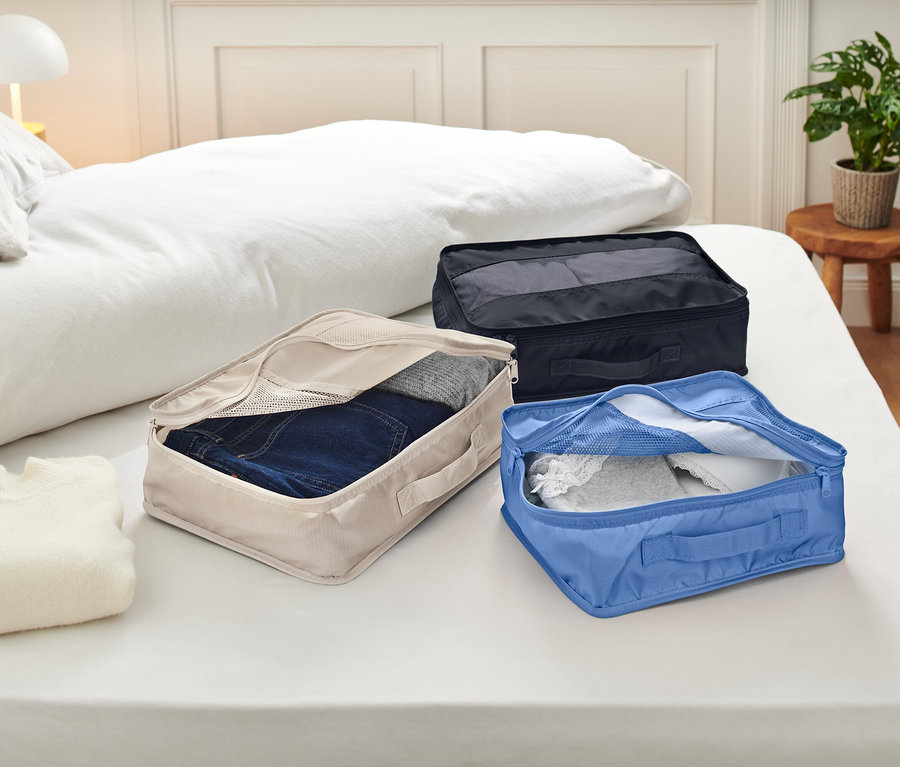 Na posteli s bílým ložním prádlem leží tři otevřené balicí kostky. Kostky jsou béžové, modré a černé a obsahují oblečení.