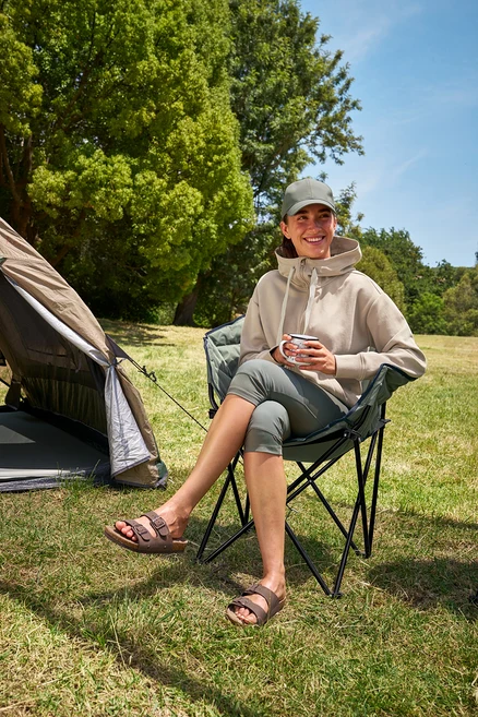 Žena sedí v kempingové židli Tambu »ASANA«, drží šálek a má na sobě 3/4 outdoorové legíny a mikinu s kapucí.