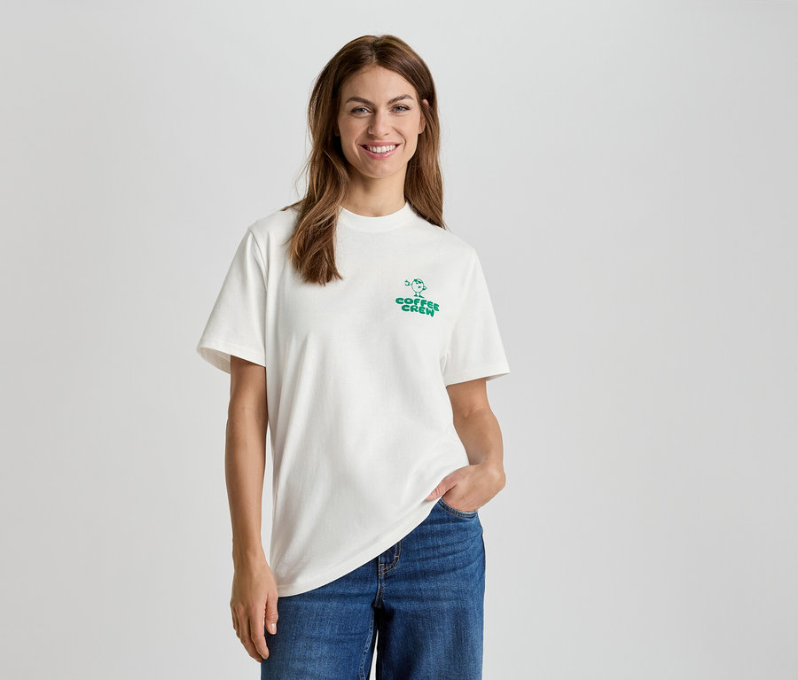 Žena v bílém tričku s nápisem "Coffee Crew" a modrých džínách.