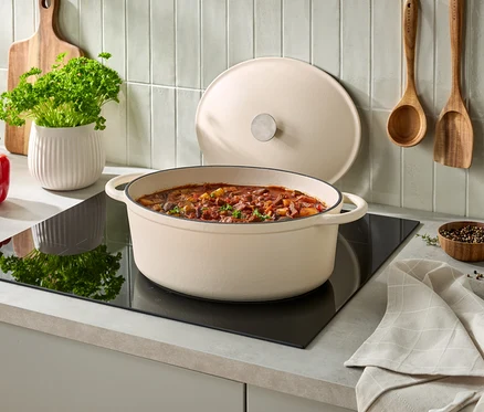 Krémový oválný litinový hrnec s gulášem na černé indukční varné desce, jeho poklice je opřená vzadu. Vlevo je červená paprika, bílý květináč s petrželkou a dřevěné prkénko. Na obložené stěně visí dva dřevěné kuchyňské nástroje, vpravo malá miska s pepřem a složená kuchyňská utěrka.