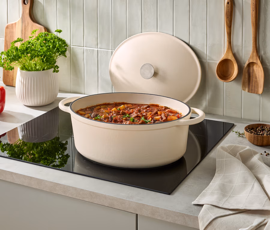 Krémový oválný litinový hrnec s gulášem na černé indukční varné desce, jeho poklice je opřená vzadu. Vlevo je červená paprika, bílý květináč s petrželkou a dřevěné prkénko. Na obložené stěně visí dva dřevěné kuchyňské nástroje, vpravo malá miska s pepřem a složená kuchyňská utěrka.