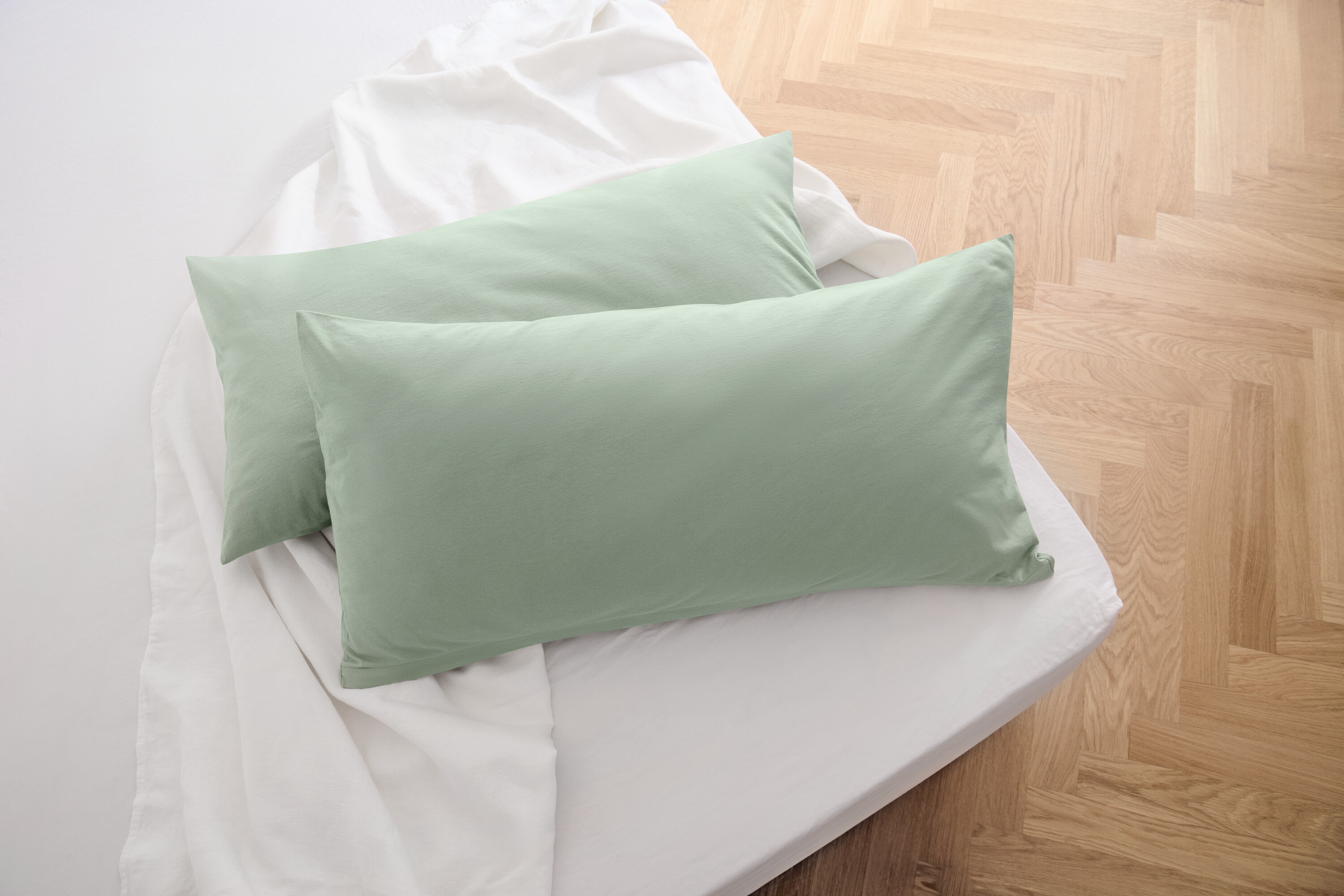 Dva žerzejové povlaky na polštáře šalvějově zelené barvy leží na bílém prostěradle.