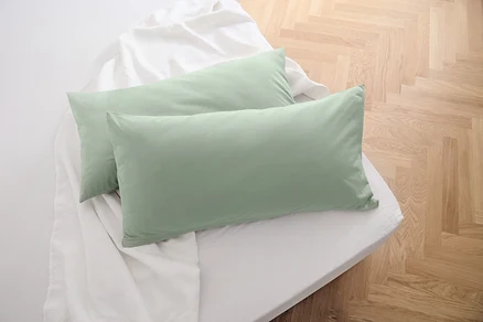 Dva žerzejové povlaky na polštáře šalvějově zelené barvy leží na bílém prostěradle.