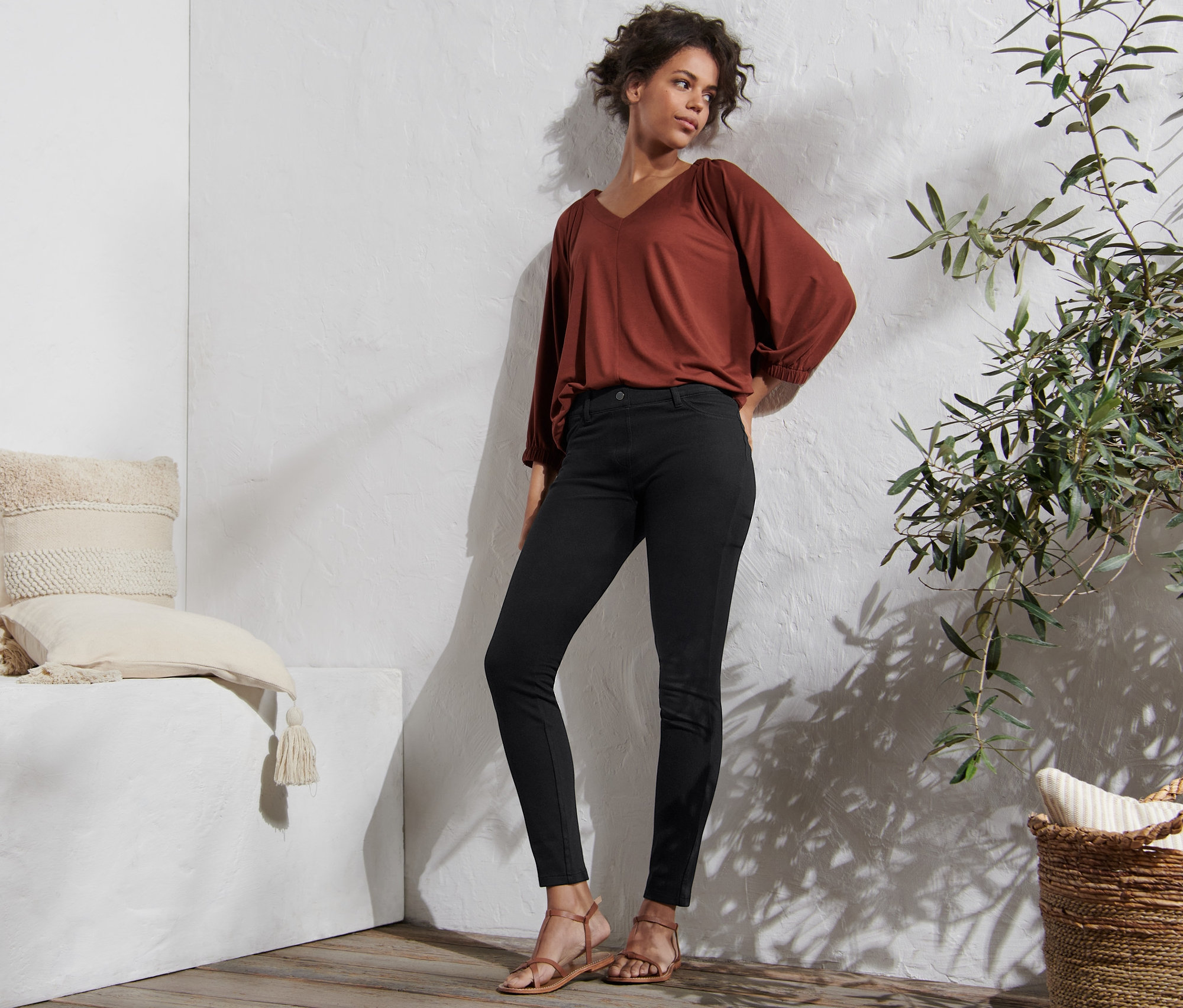 Žena pózuje v černých džegínách, hnědé halence a sandálech.