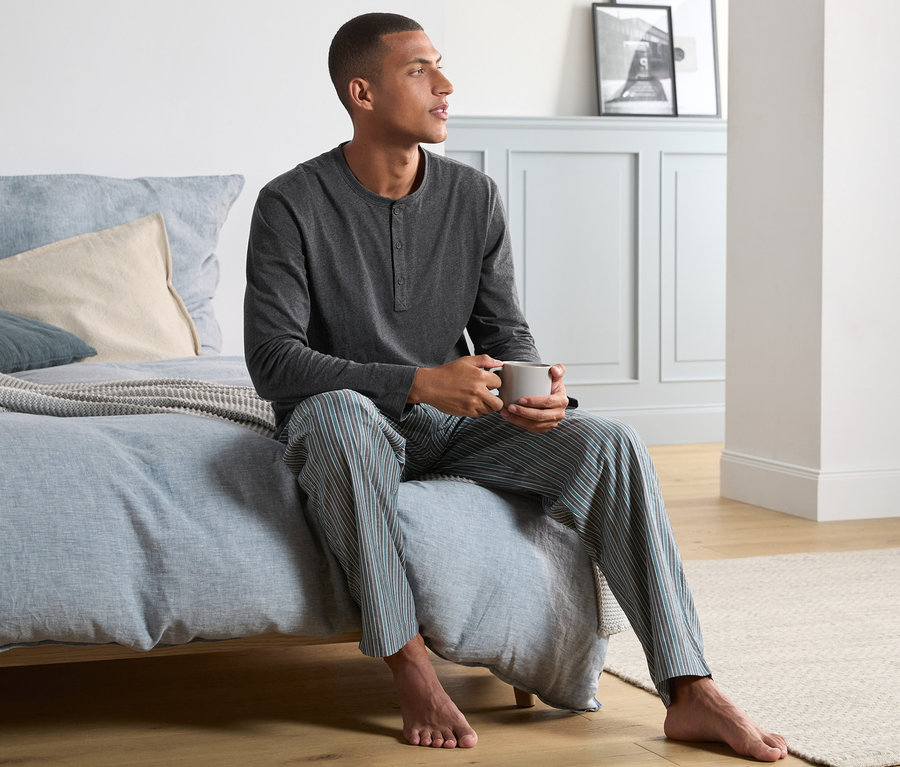 Muž sedí na posteli a pije z hrnku. Má na sobě pyžamo s tkanými kalhotami.