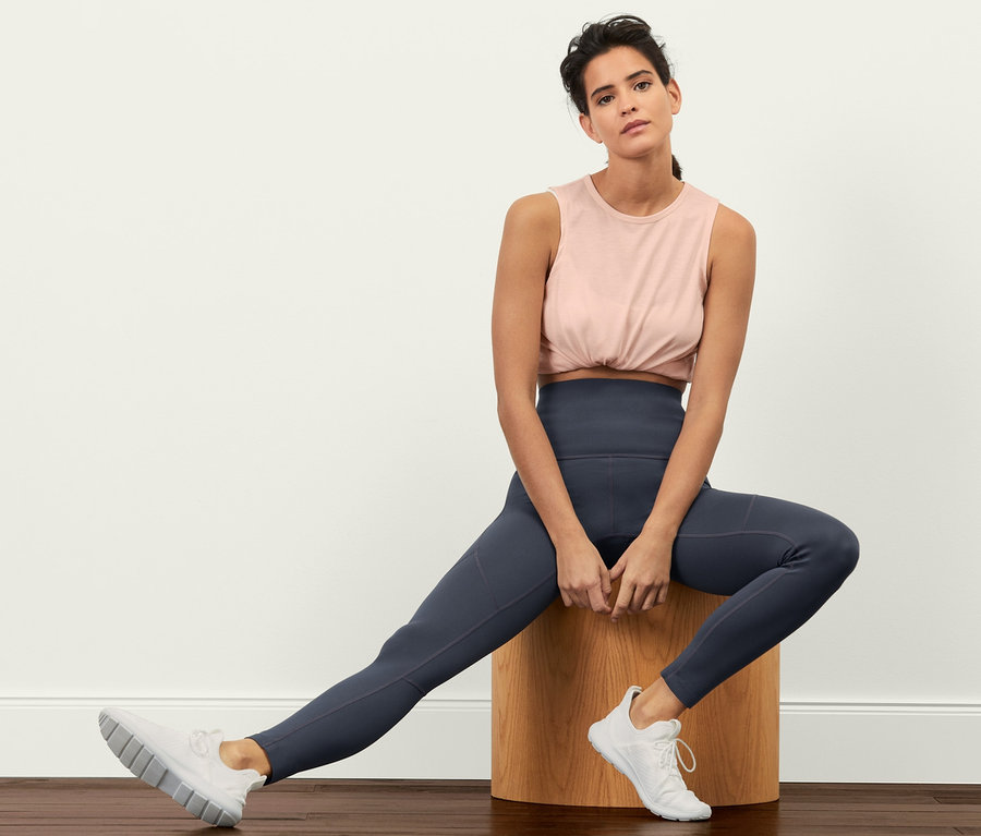 Žena sedí na dřevěné stoličce a má na sobě sportovní legíny Sculpture, top a tenisky.