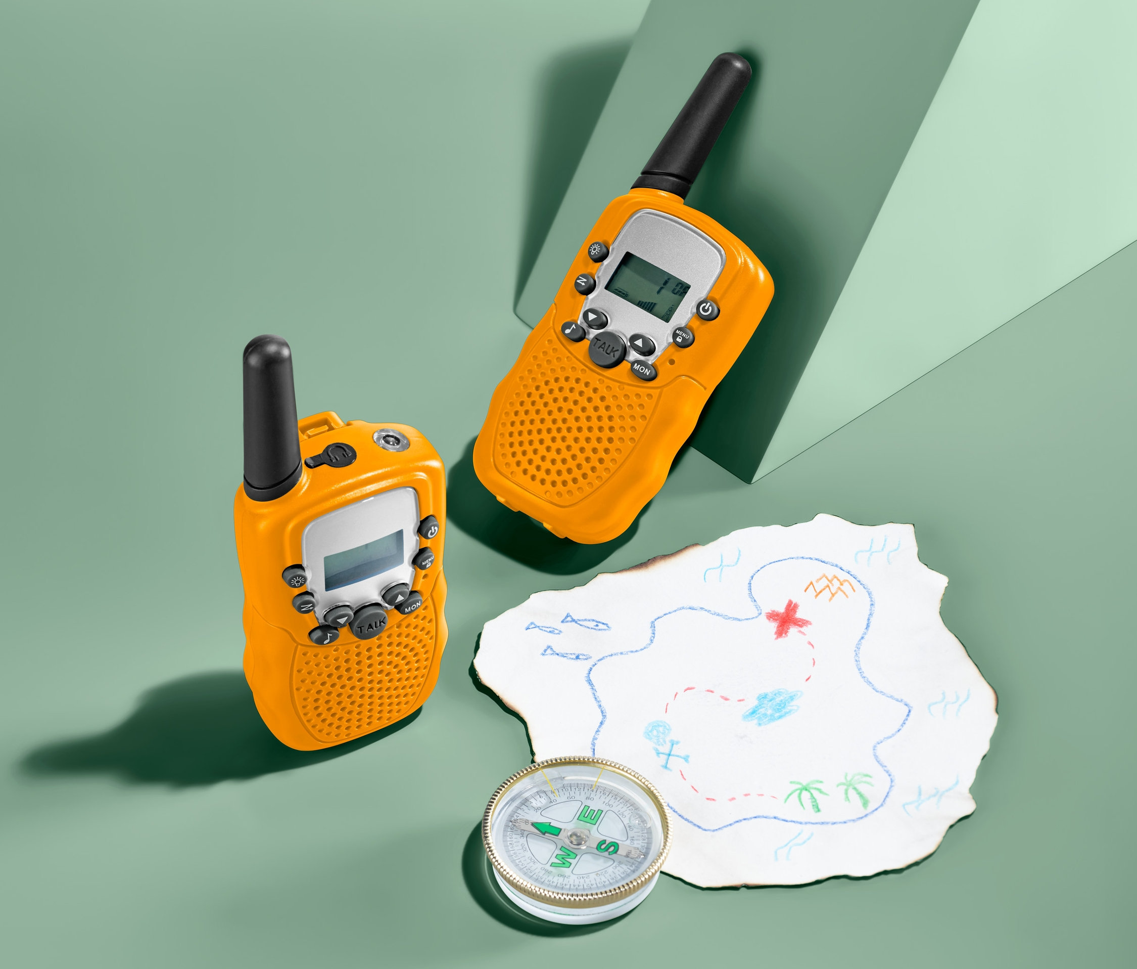 Dvě dětské vysílačky Walkie Talkie a kompas leží vedle mapy pokladu na zeleném pozadí.