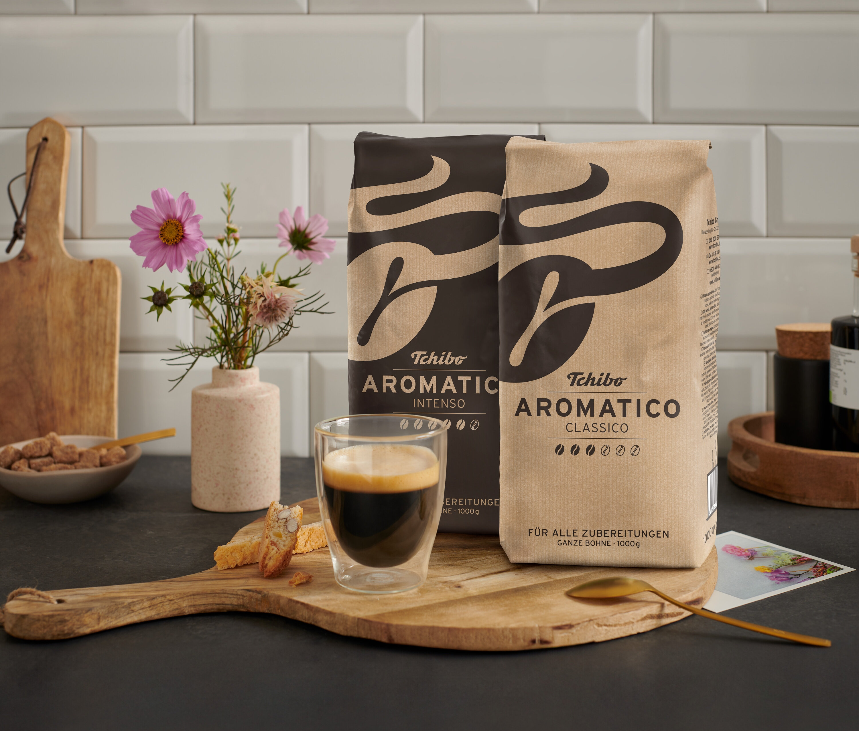 Dvě balení kávy Tchibo Aromatica a šálek espressa na dřevěném stole s květinami a pečivem.