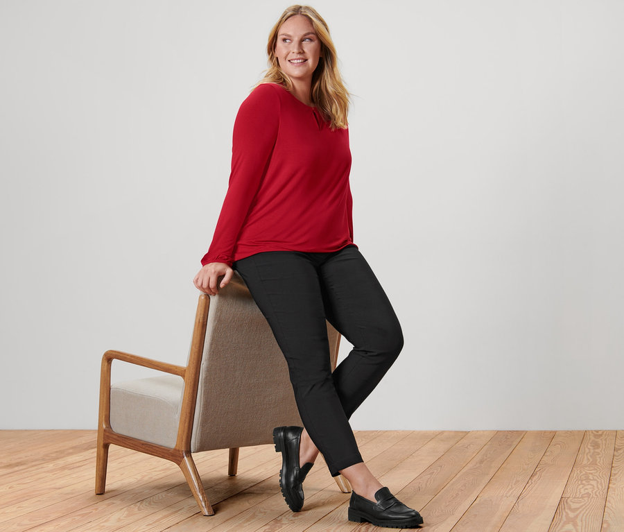 Žena sedí na křesle a má na sobě červené halenkové triko, černé bengalínové kalhoty a kožené mokasíny.