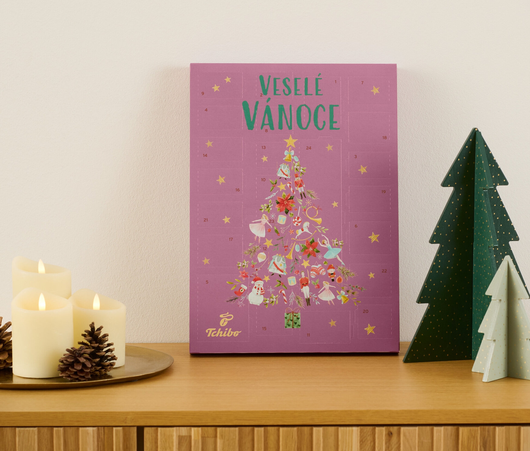 Adventní kalendář Tchibo na dřevěné komodě vedle svíček a ozdobných stromků.