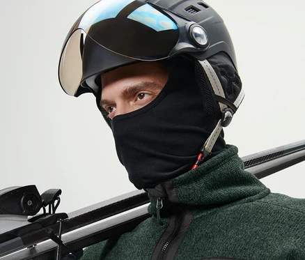 Muž v lyžařské helmě a kukle Performance nese lyže přes rameno.