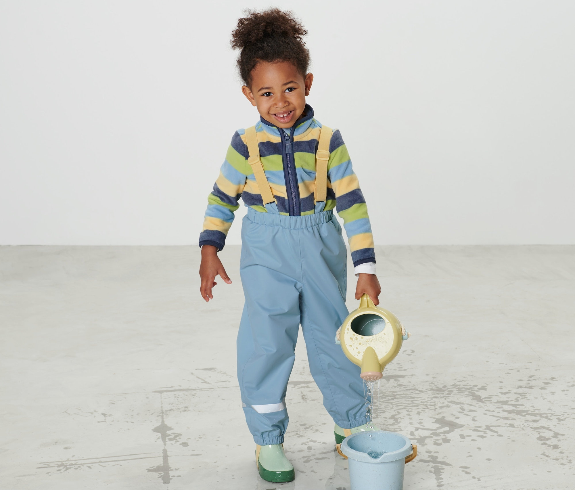 Chlapec v dětských kalhotách do deště lije vodu z konev do kyblíku.