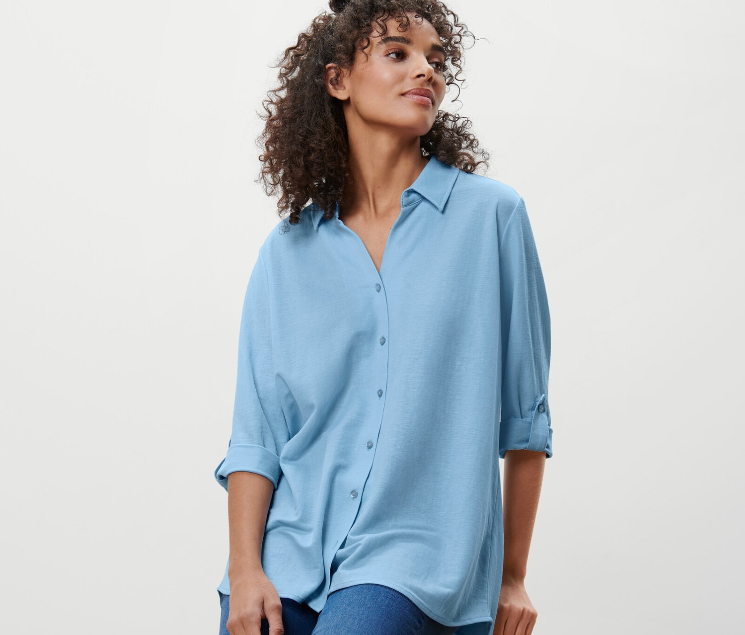 Žena sedí na stoličce a má na sobě světle modrou halenku z žerzejového piké a džíny.