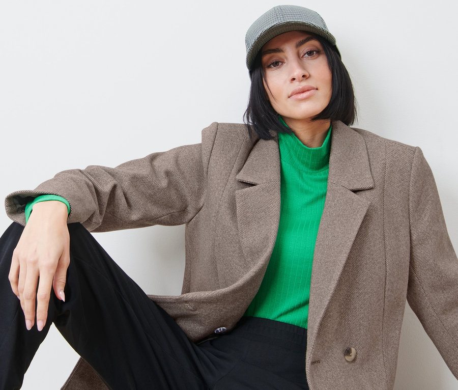 Žena sedí a má na sobě blejzr, zelený rolák a kšiltovku.