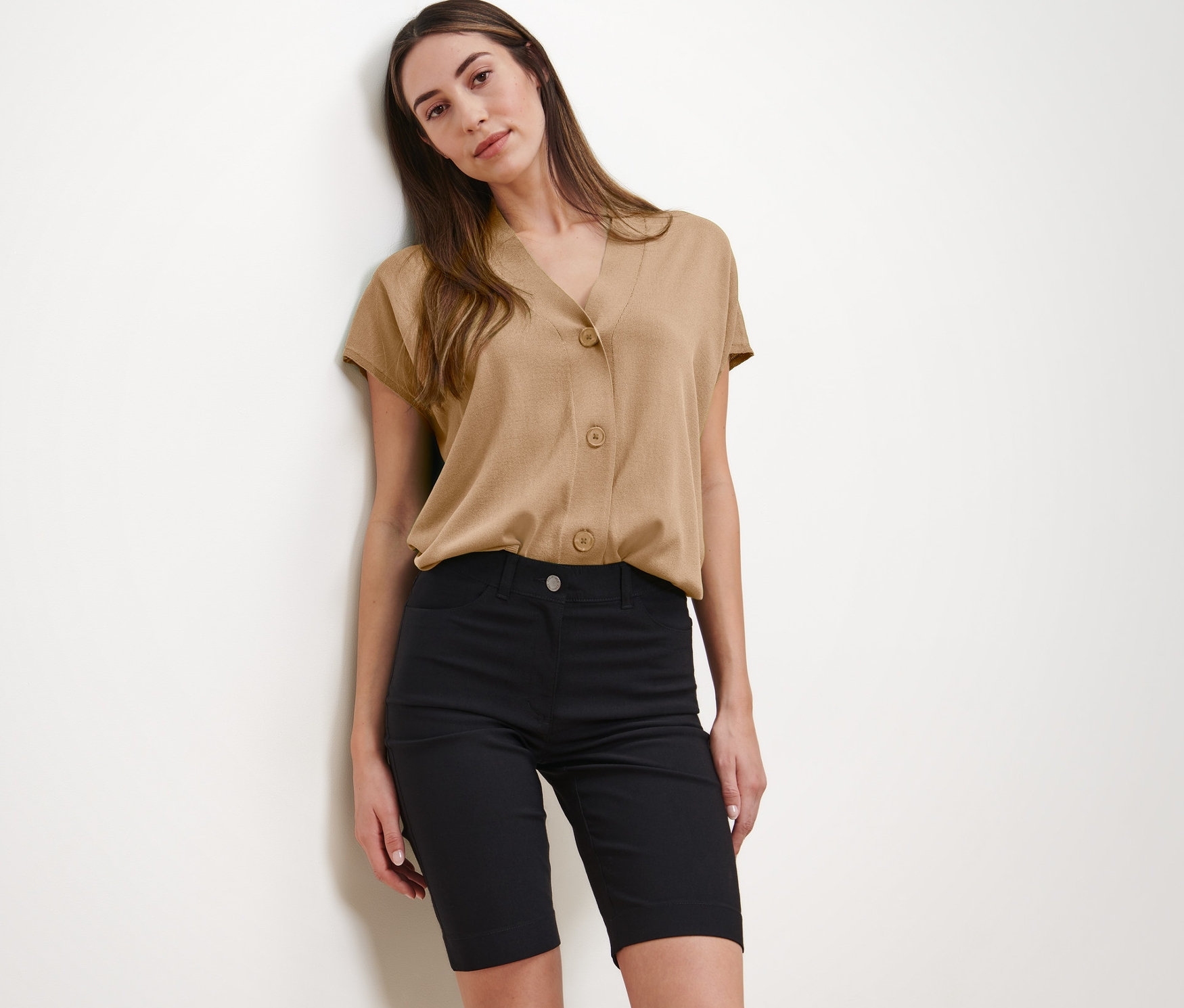 Žena pózuje v béžové halence a černých bermudách.