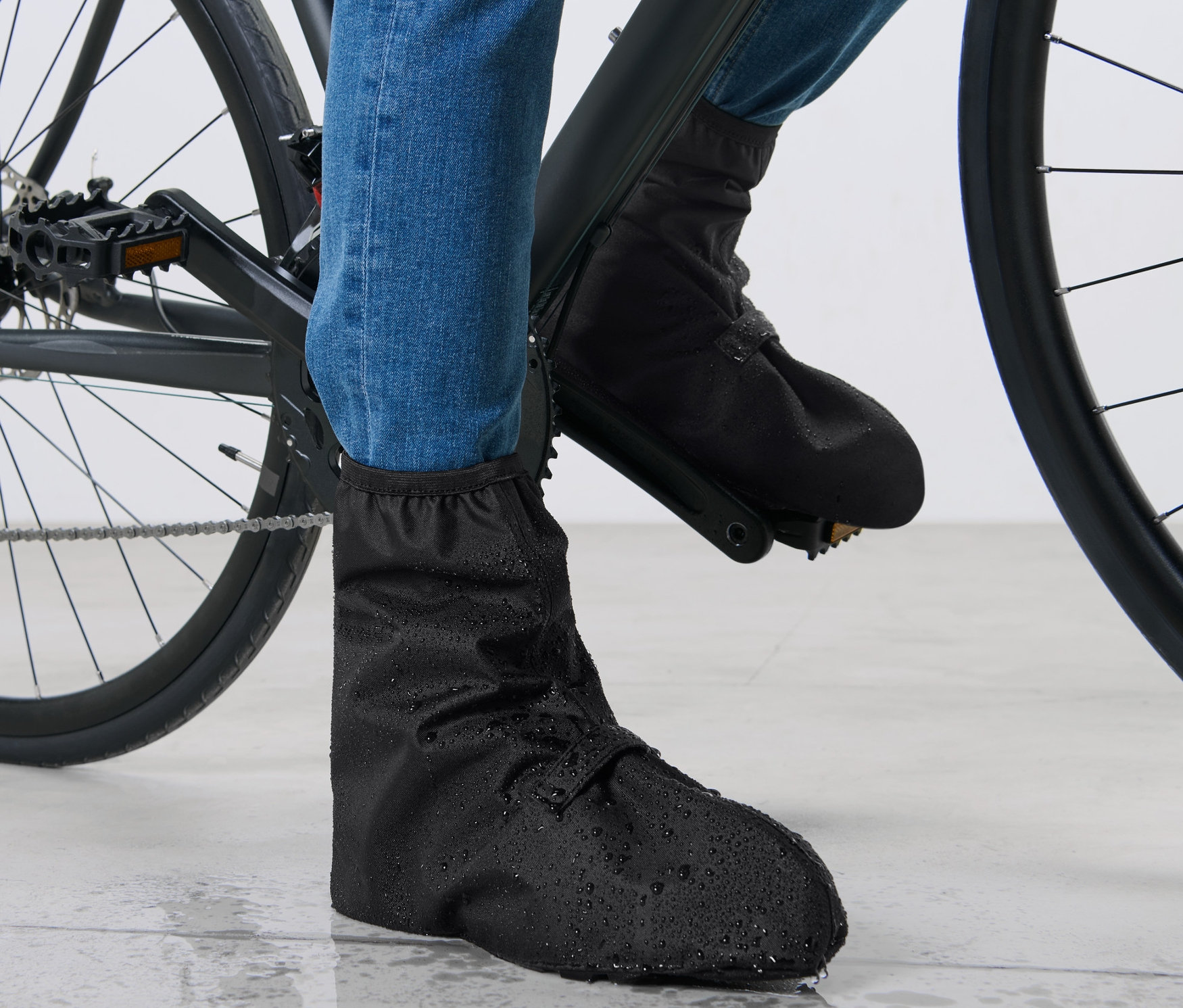 Nohy cyklisty v modrých džínách a černých návlecích na boty do deště na kole.