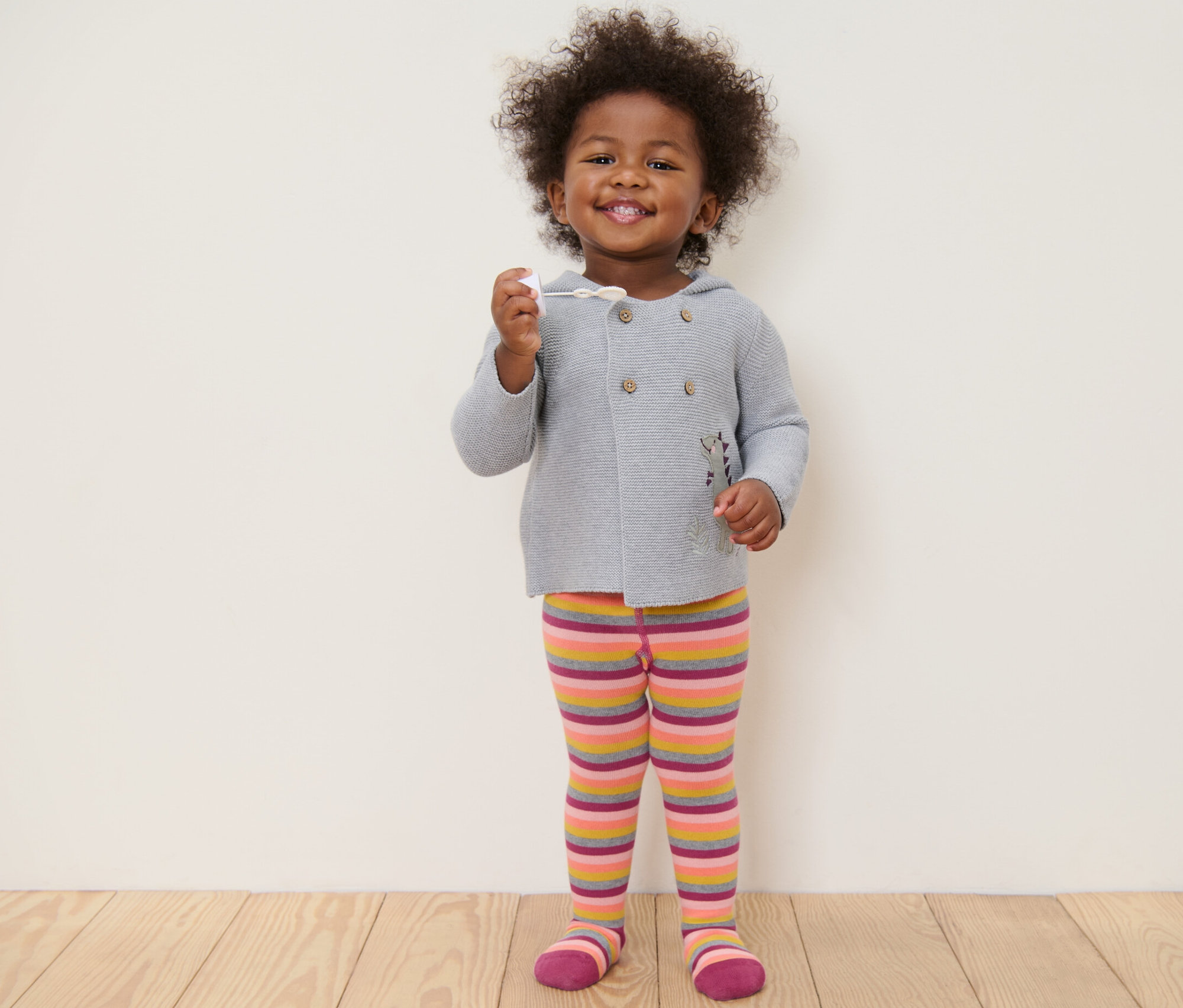 Usmívající se dítě stojí a drží v ruce hračku. Má na sobě šedý svetr a růžové pruhované punčocháče.