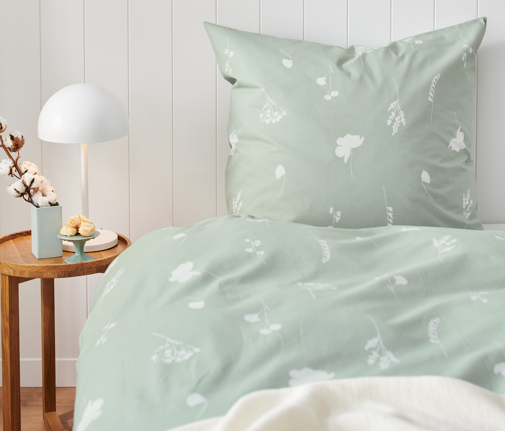 Detailní záběr na ložnici s mátově zeleným povlečením s květinovým vzorem, nočním stolkem a lampou.