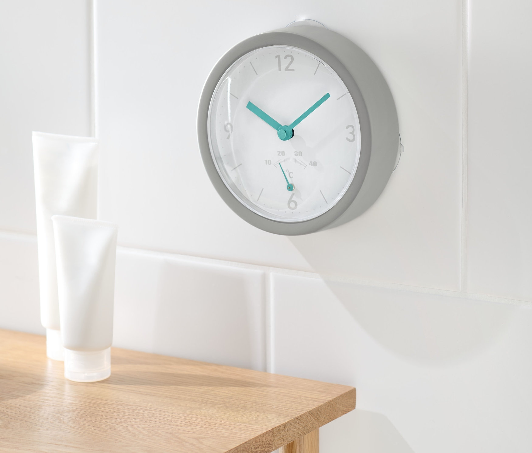Na bílé zdi koupelny visí bezdrátové nástěnné hodiny s teploměrem. Na dřevěném stolku stojí dvě bílé tuby.