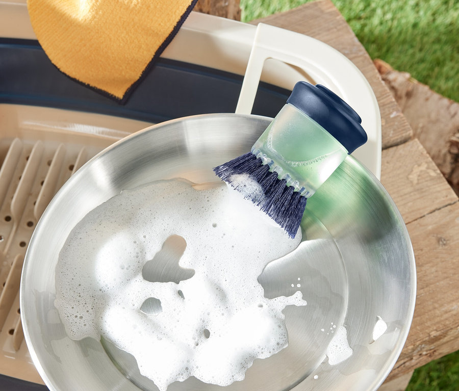 Nerezová pánev je umývána kartáčem na mytí nádobí s dávkovačem saponátu. Vedle je odkapávač a hadr.