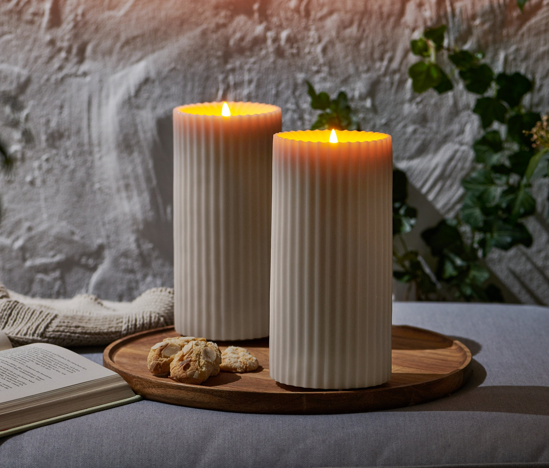 Dvě LED svíčky z pravého vosku do exteriéru na dřevěném podnose s knihou a sušenkami.