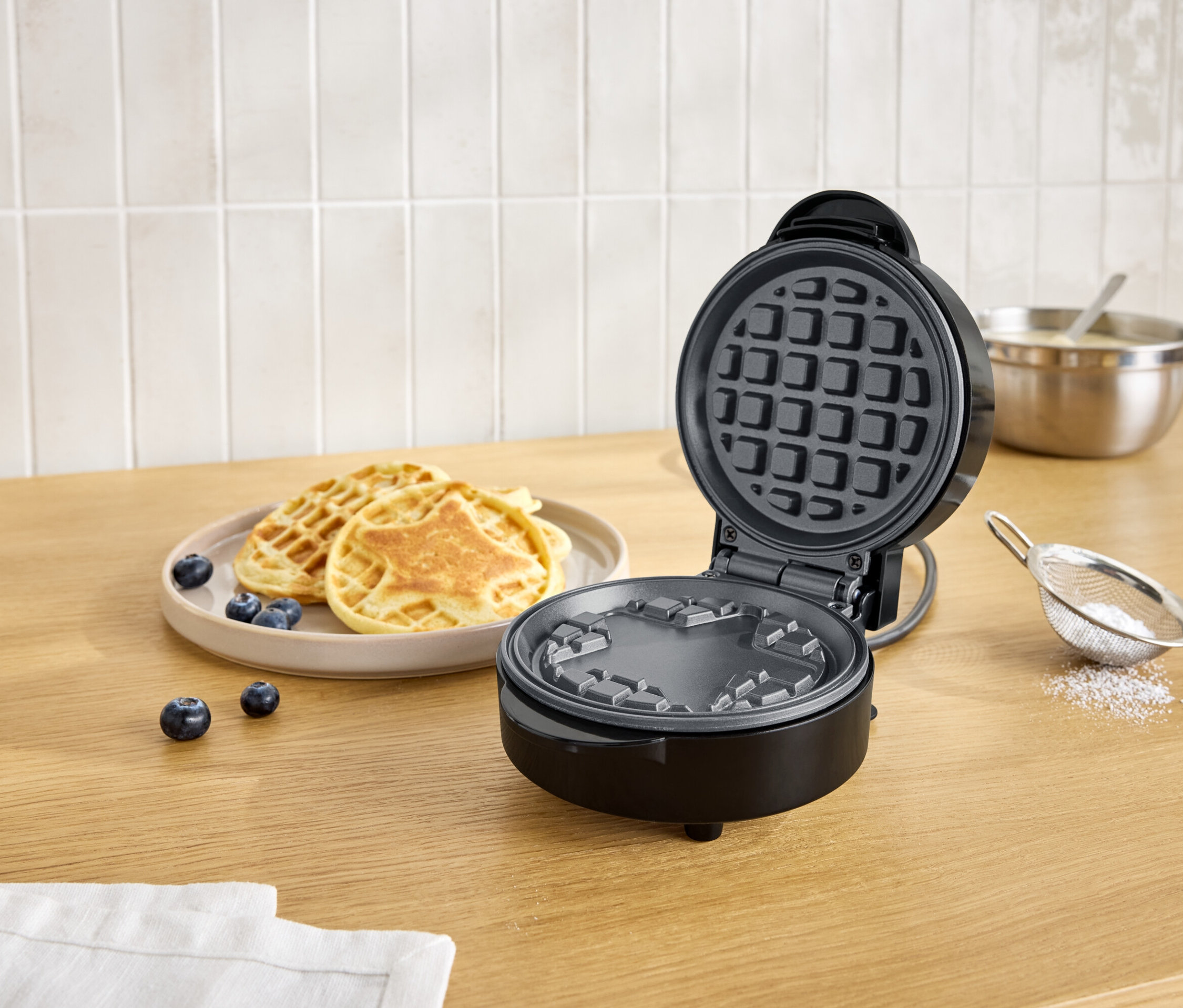 Otevřený černý vaflovač vedle talíře s vaflemi a borůvkami na dřevěném stole.
