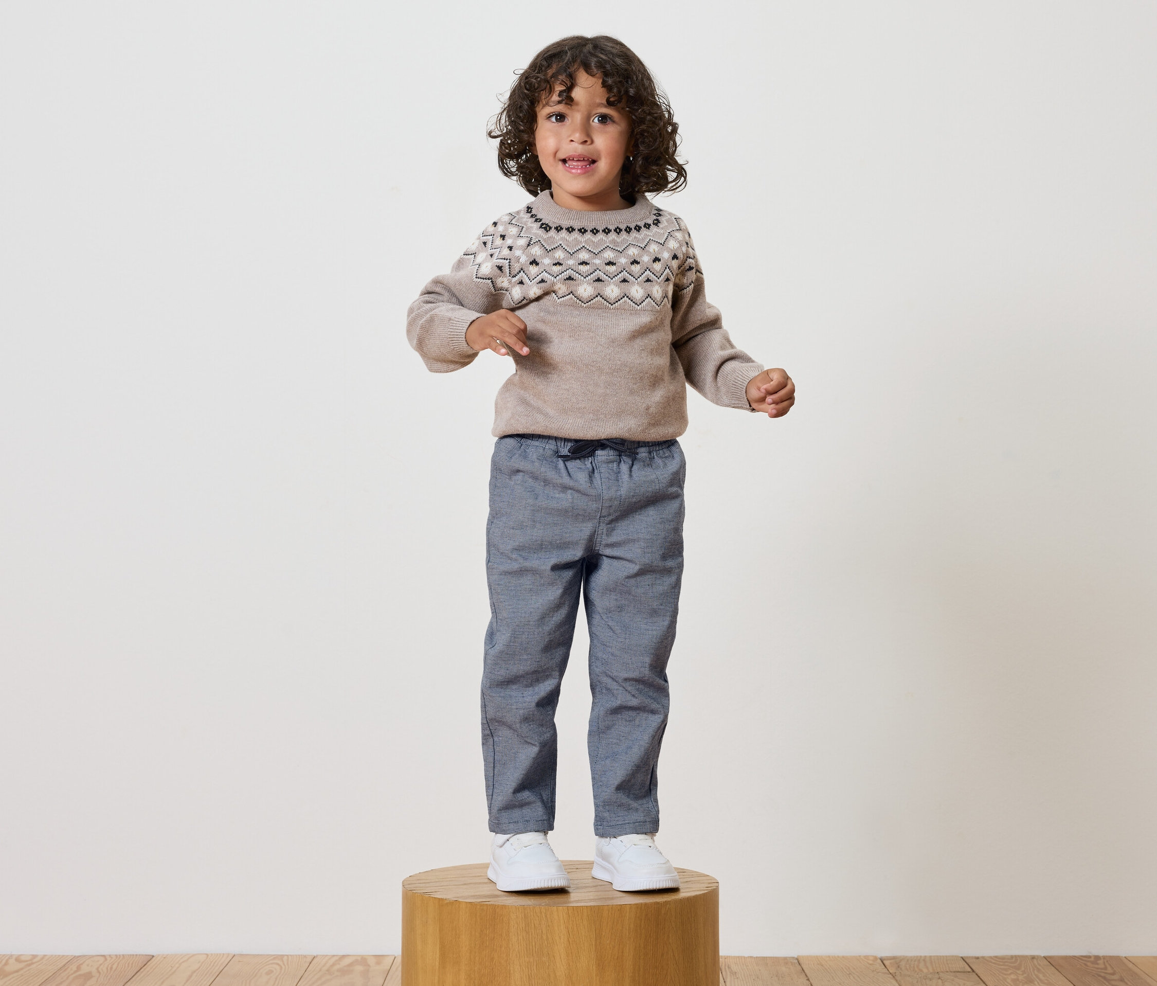 Chlapec s kudrnatými vlasy stojí na dřevěném podstavci, má na sobě svetr se vzorem a modré kalhoty.