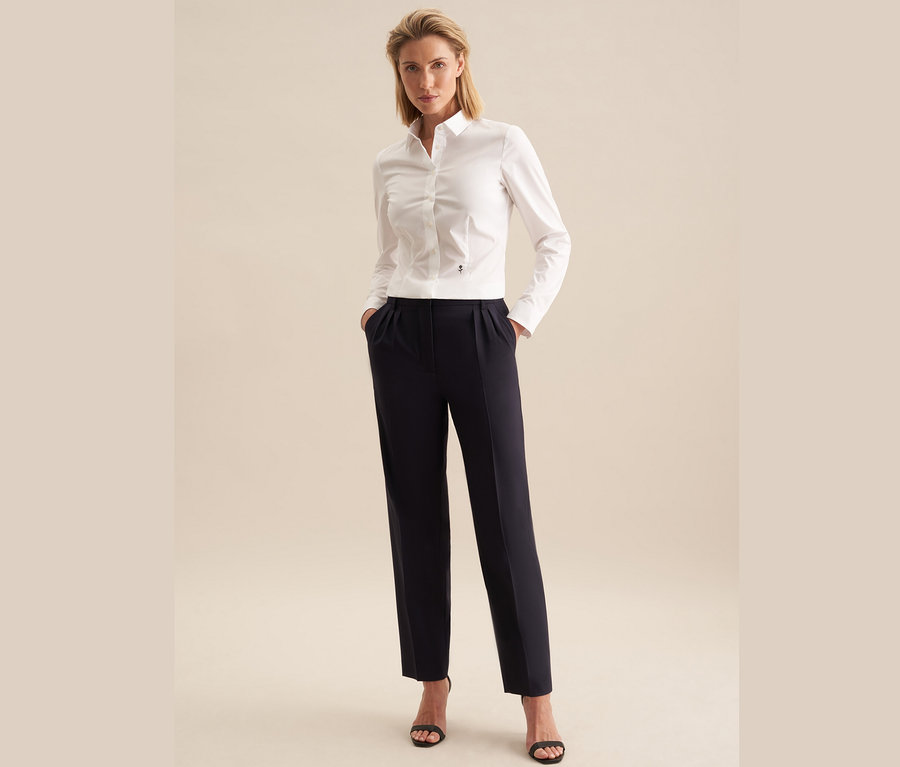 Žena stojí a má ruce v kapsách kalhot. Má na sobě bílou halenku slim fit Seidensticker, tmavě modré kalhoty a černé sandály.