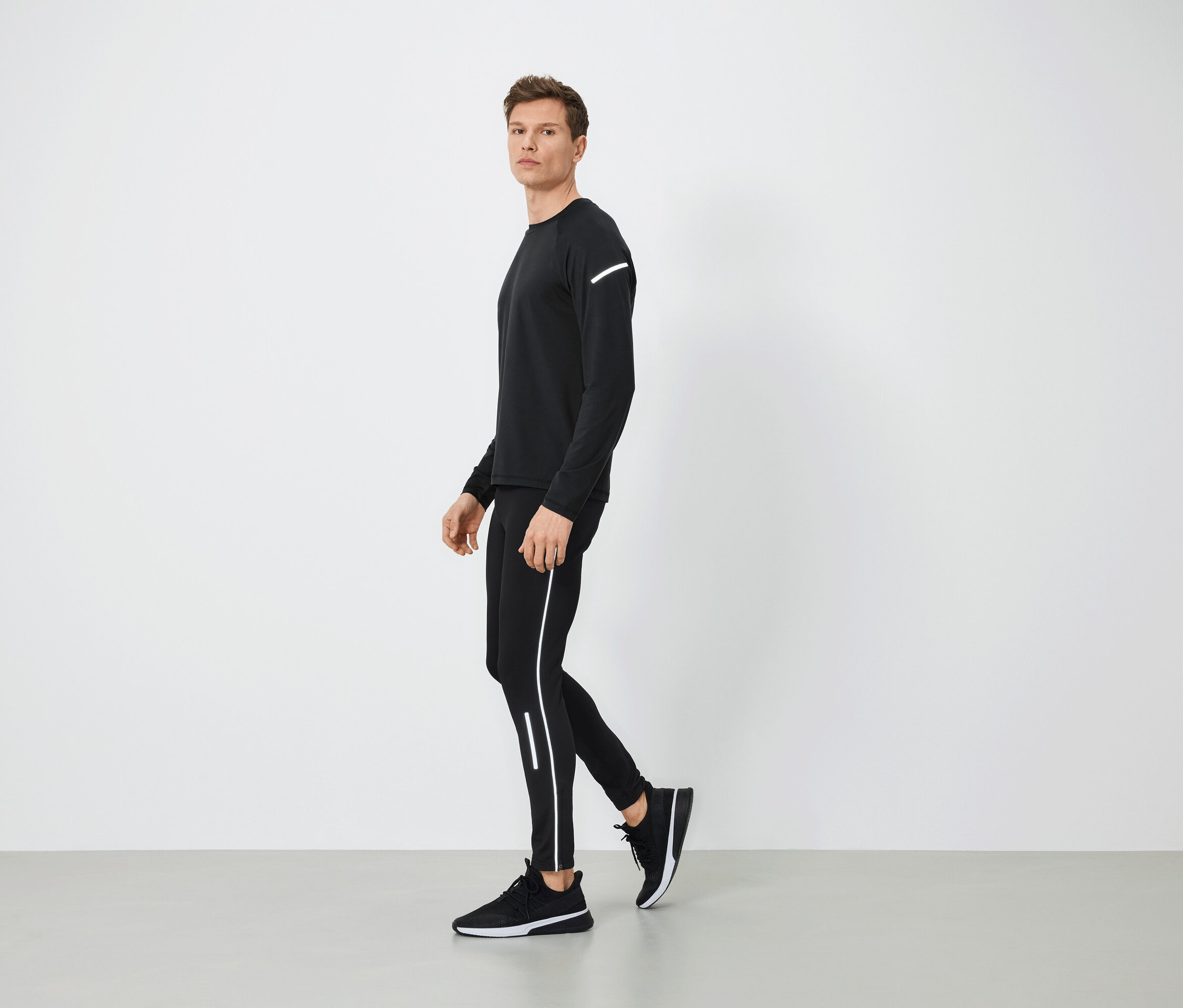 Muž v černém sportovním oblečení s reflexními prvky kráčí po šedé podlaze.