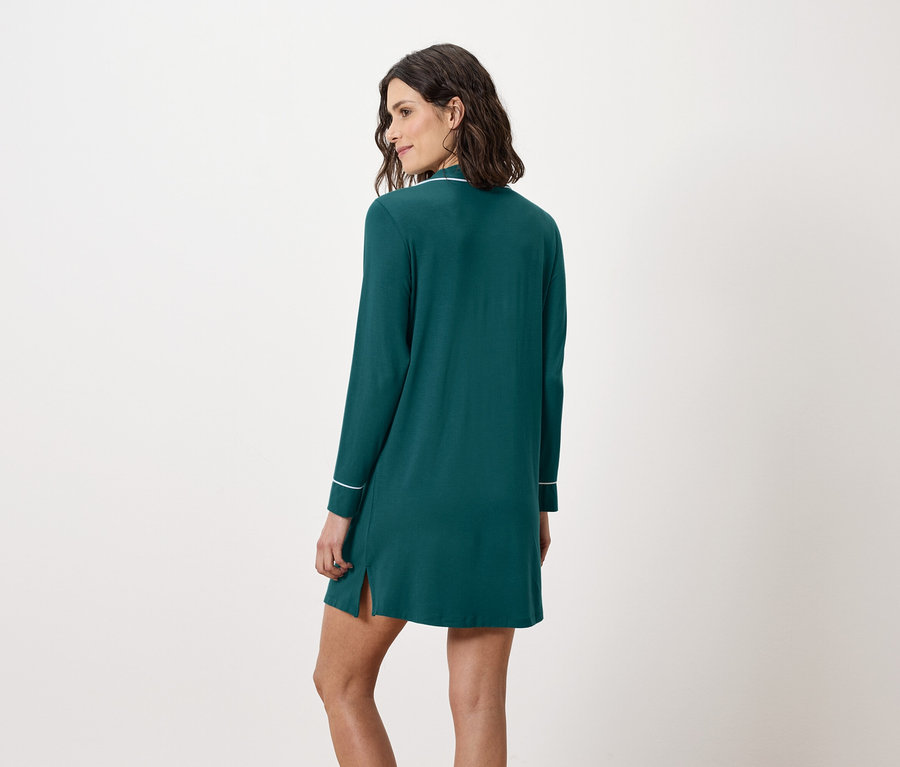 Žena stojí zády a má na sobě smaragdově zelenou noční košili s dlouhými rukávy a bílým lemováním.