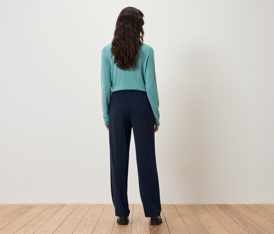 Zezadu je vidět žena s tmavě hnědými vlasy v tyrkysovém svetru a tmavě modrých kalhotách.