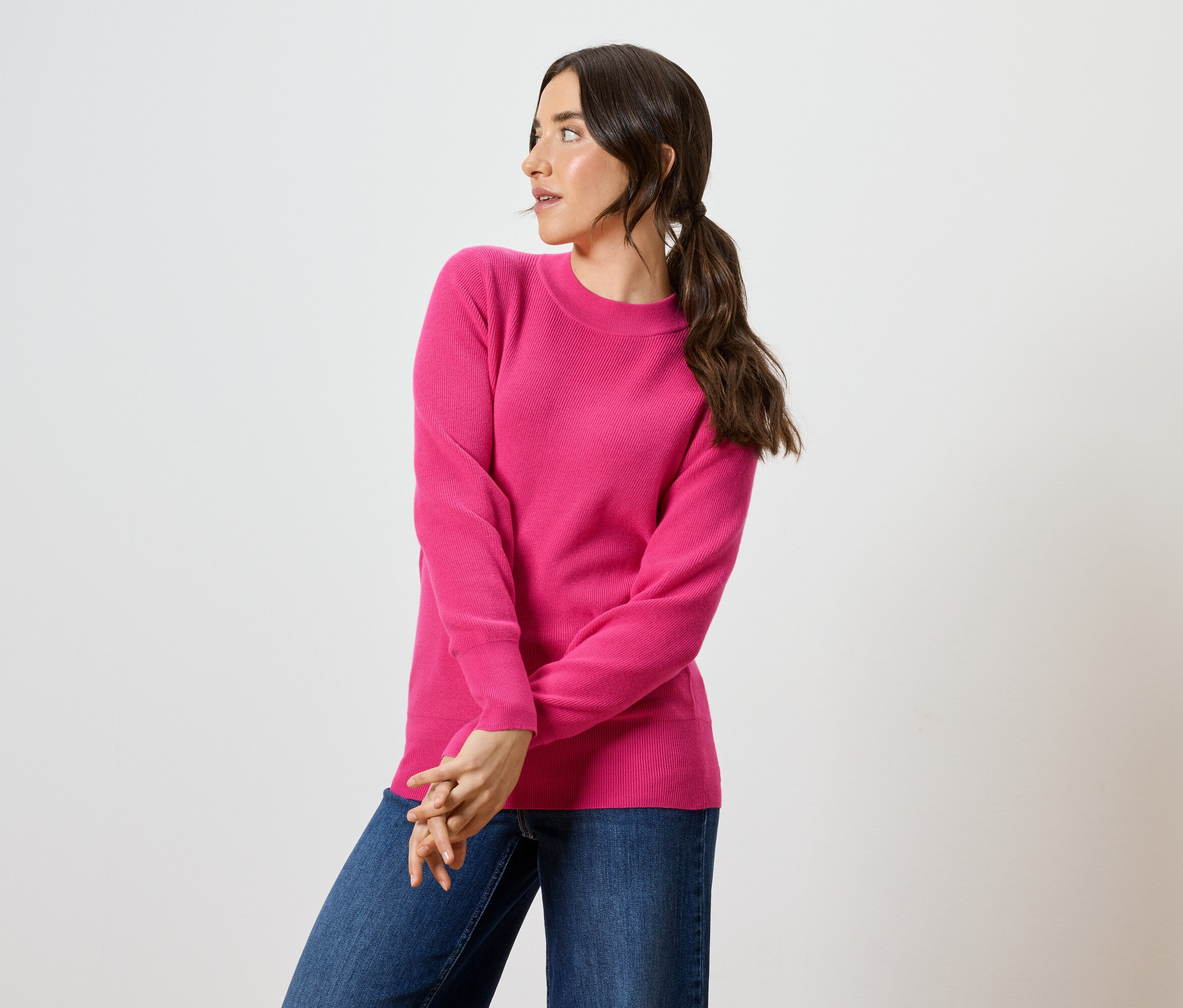 Žena s hnědými vlasy a modrýma očima pózuje v růžovém svetru a modrých džínách.