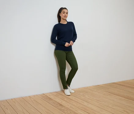 Žena stojí opřená o zeď a má na sobě tmavě modrý svetr, zelené džíny a bílé tenisky.