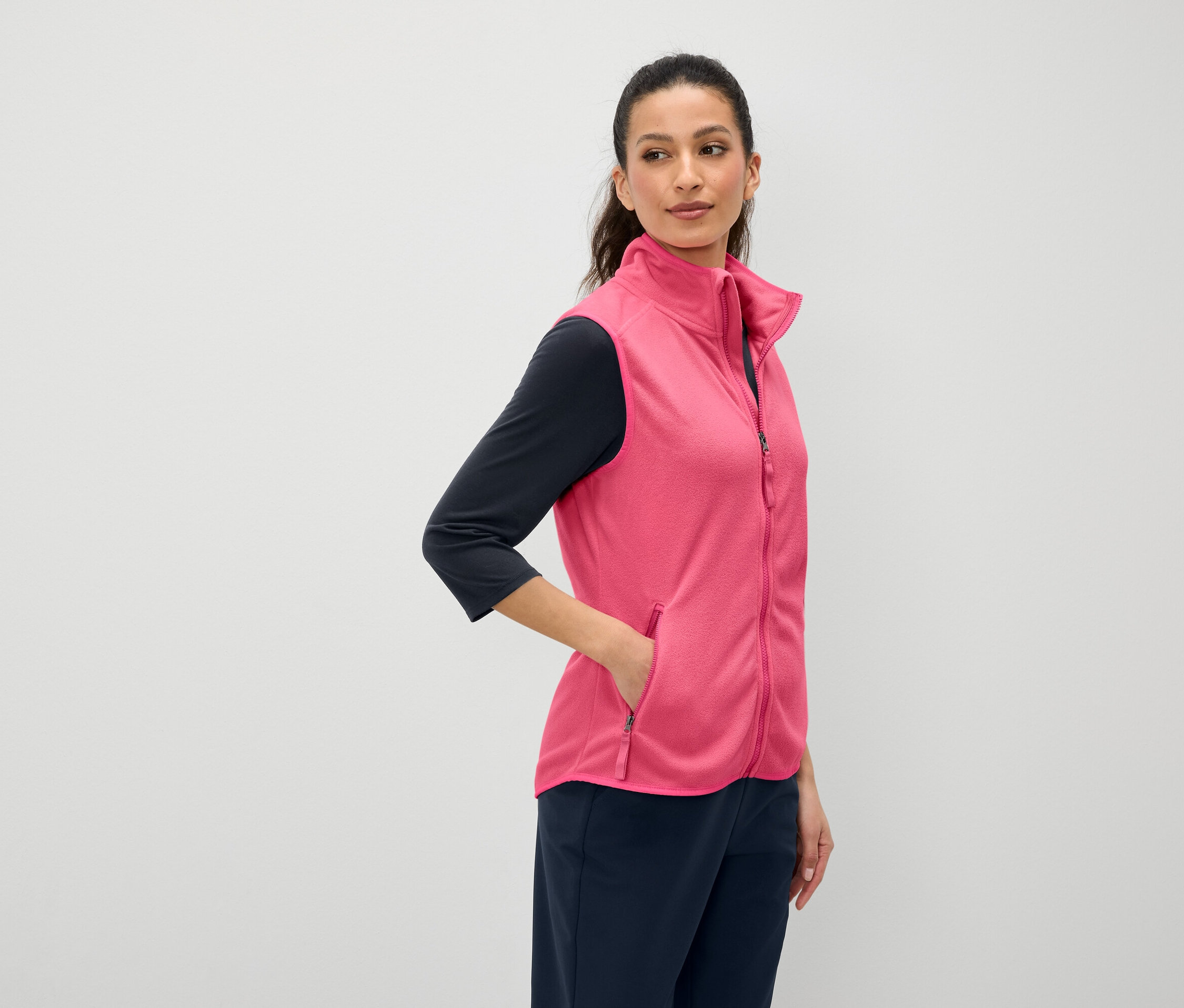 Žena s hnědými vlasy má ruku v kapse růžové vesty. Oblečená je do tmavomodrého trička s tříčtvrtečním rukávem a tmavomodrých kalhot.