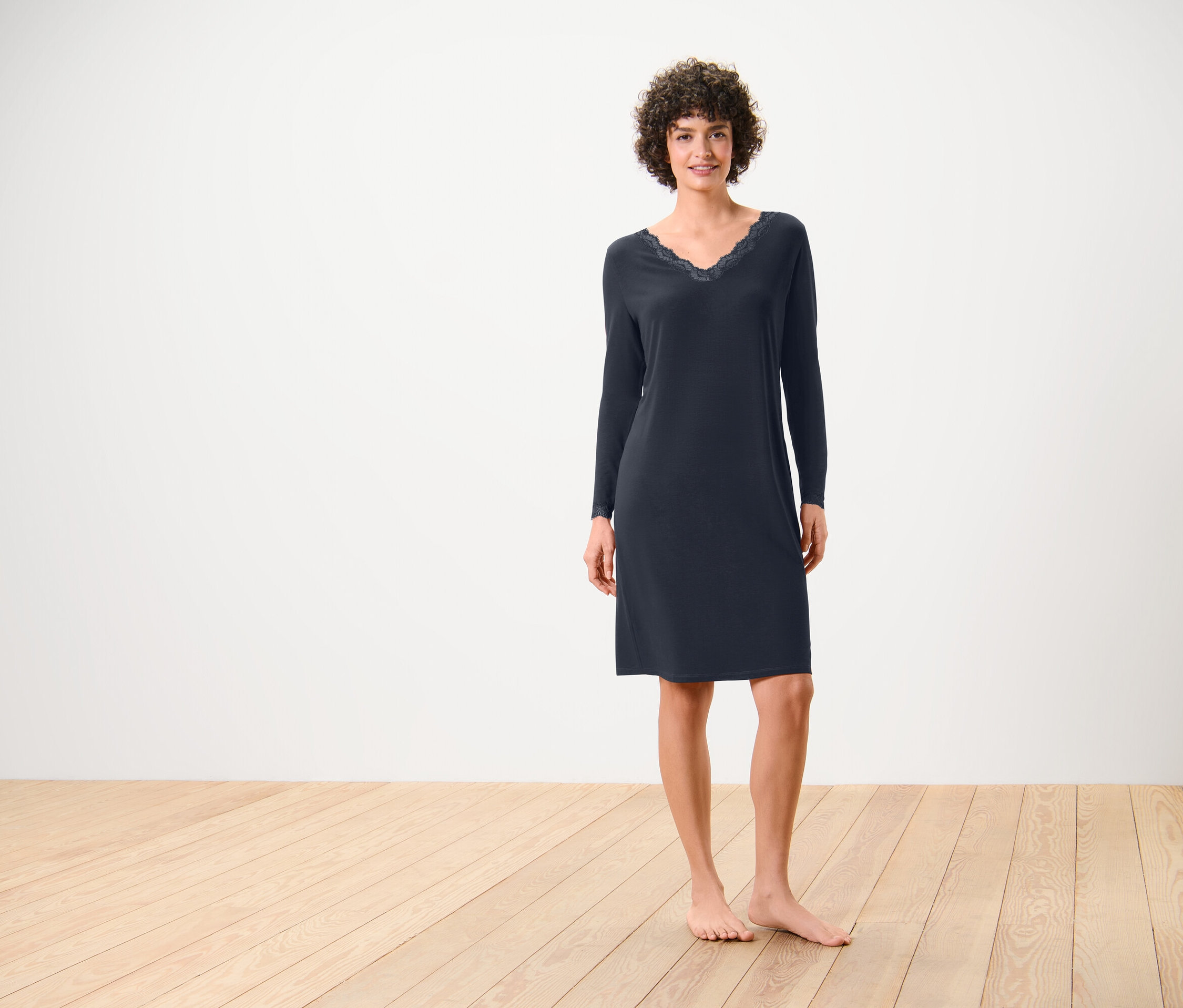 Žena stojí na dřevěné podlaze a má na sobě tmavě modré noční šaty s dlouhými rukávy a krajkou na výstřihu.