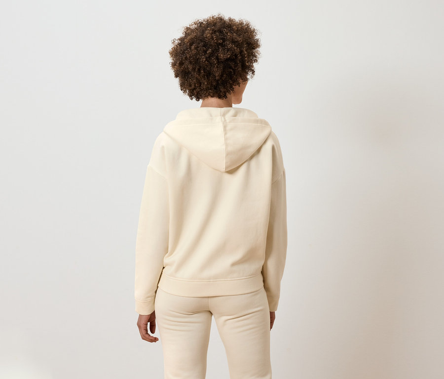 Zezadu je vidět žena s kudrnatými vlasy v béžové soupravě s kapucí.