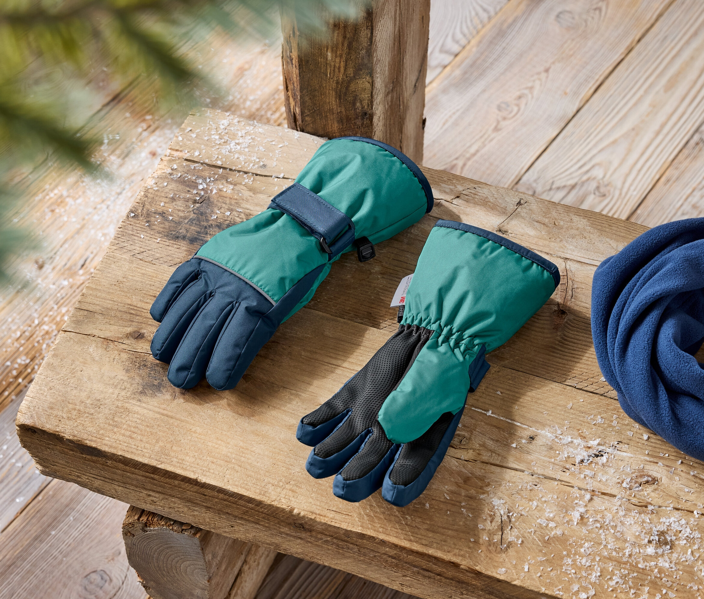 Zimní rukavice v zelené a tmavě modré barvě a modrá fleecová šála leží na dřevěné lavici.