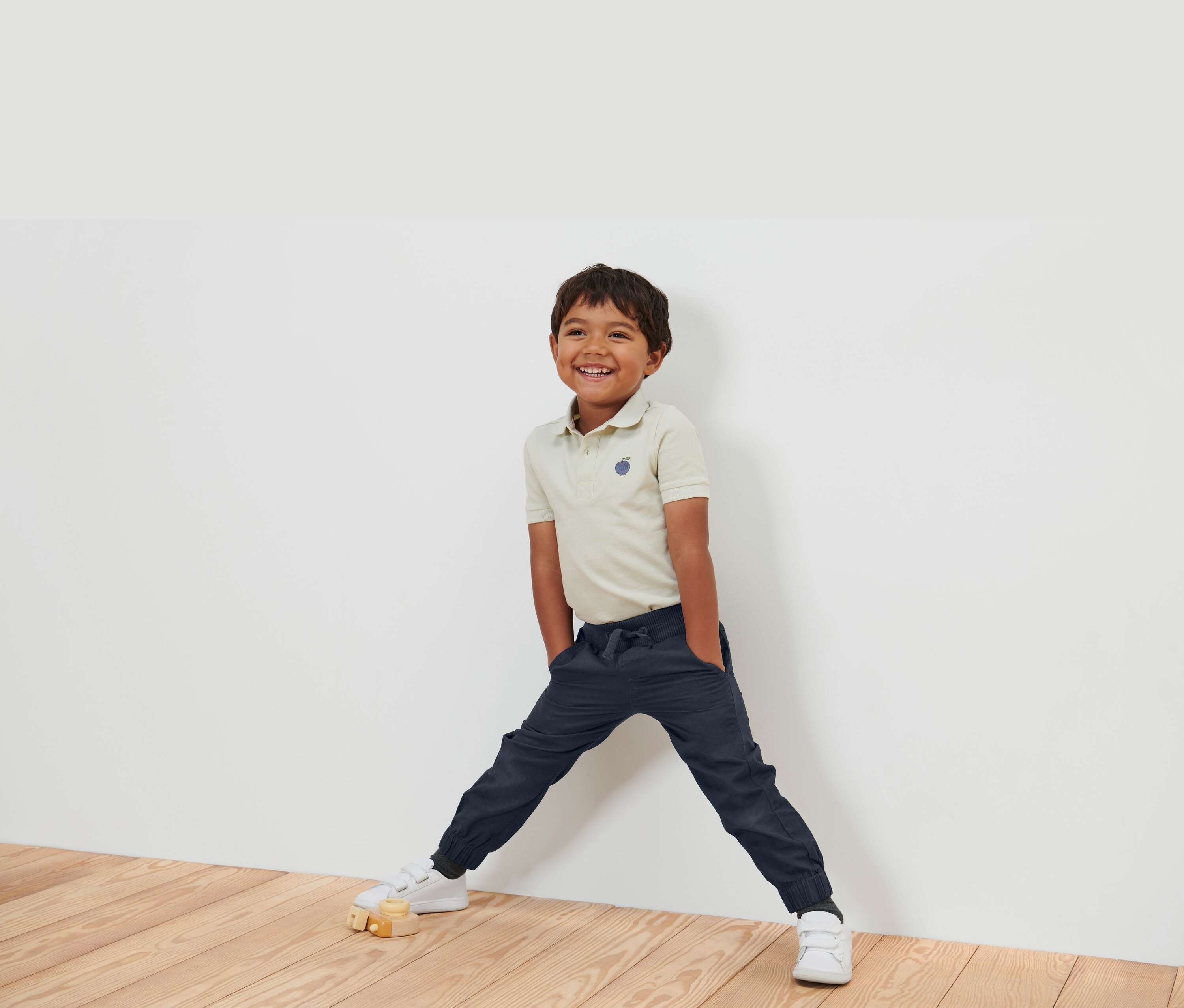 Usmívající se chlapec v béžové polokošili, tmavě modrých kalhotách a bílých botách stojí opřený o bílou zeď.