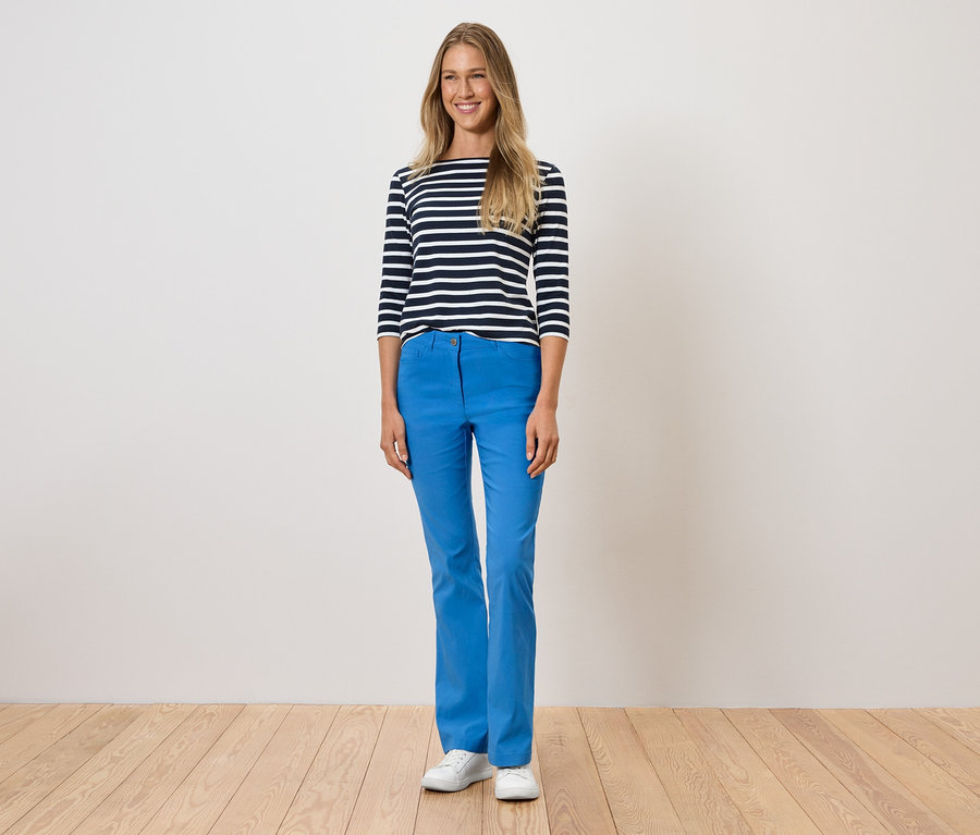 Žena stojí a má na sobě modré kalhoty, pruhované tričko a bílé boty.