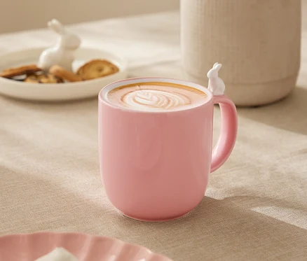 Na stole stojí růžový hrnek s bílým králíčkem na okraji, naplněný nápojem s latte art. V pozadí je bílá váza s květinami, talíř s sušenkami a figurka králíčka.