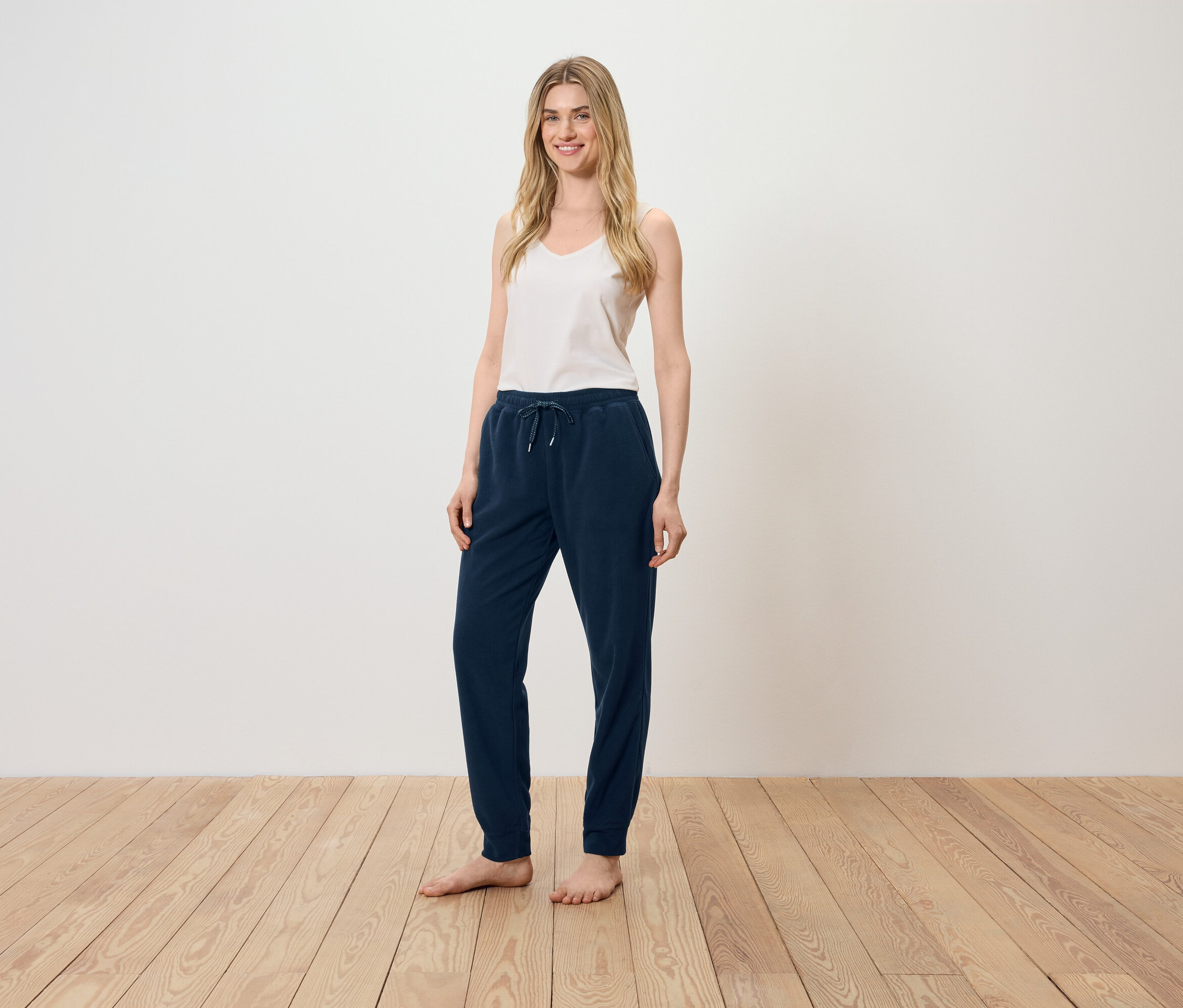 Žena stojí na dřevěné podlaze a má na sobě bílý top a tmavě modré tepláky.