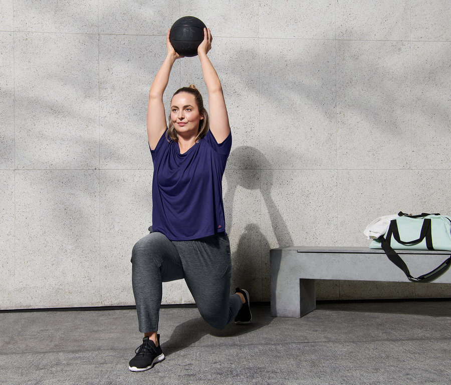 Žena dělá výpady a drží nad hlavou medicinbal, má na sobě sportovní kalhoty a fialové sportovní triko ve stylu oversized.