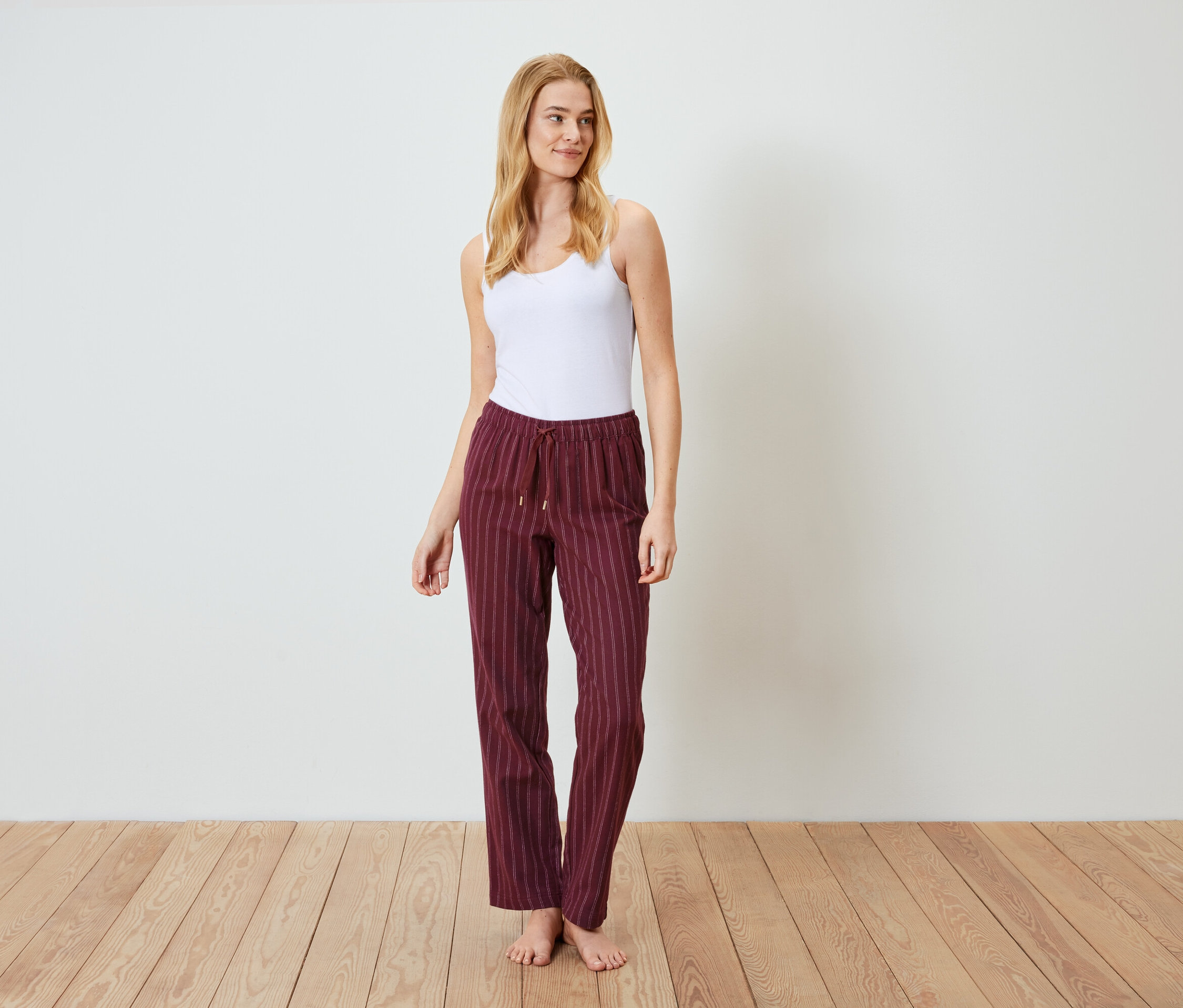 Blondýna stojí na dřevěné podlaze a má na sobě bílé tílko a vínové pruhované kalhoty.