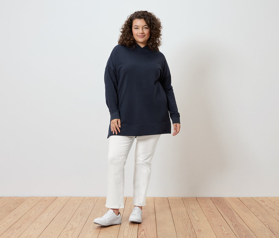 Žena s kudrnatými vlasy stojí před zdí a má na sobě tmavě modrou mikinu s kapucí, bílé džíny a bílé tenisky.