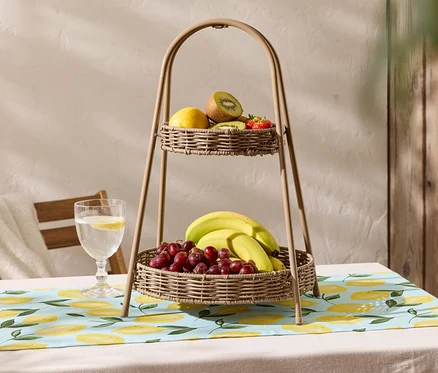 Na stole stojí dvoupatrový košík na ovoce z proutí s ubrusem s citronovým vzorem. Na horním košíku je citron, kiwi a jahody, zatímco na spodním košíku jsou banány a červené hrozny.